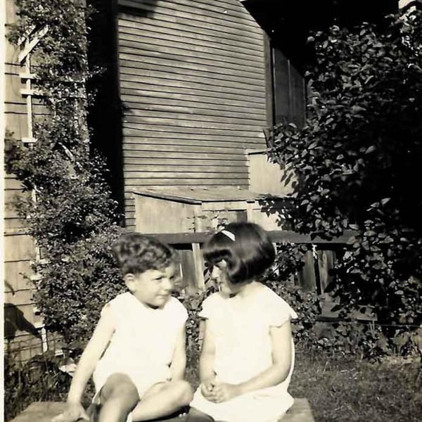 Two young children, a boy and a girl, sitting outdoors in front of a wooden building with foliage.