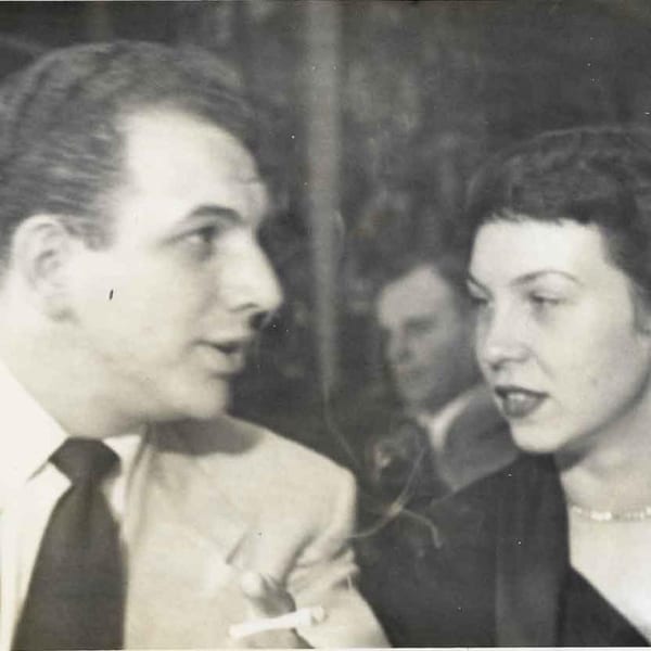 A vintage black and white photo of a man smoking and a woman talking, with another person in the background.