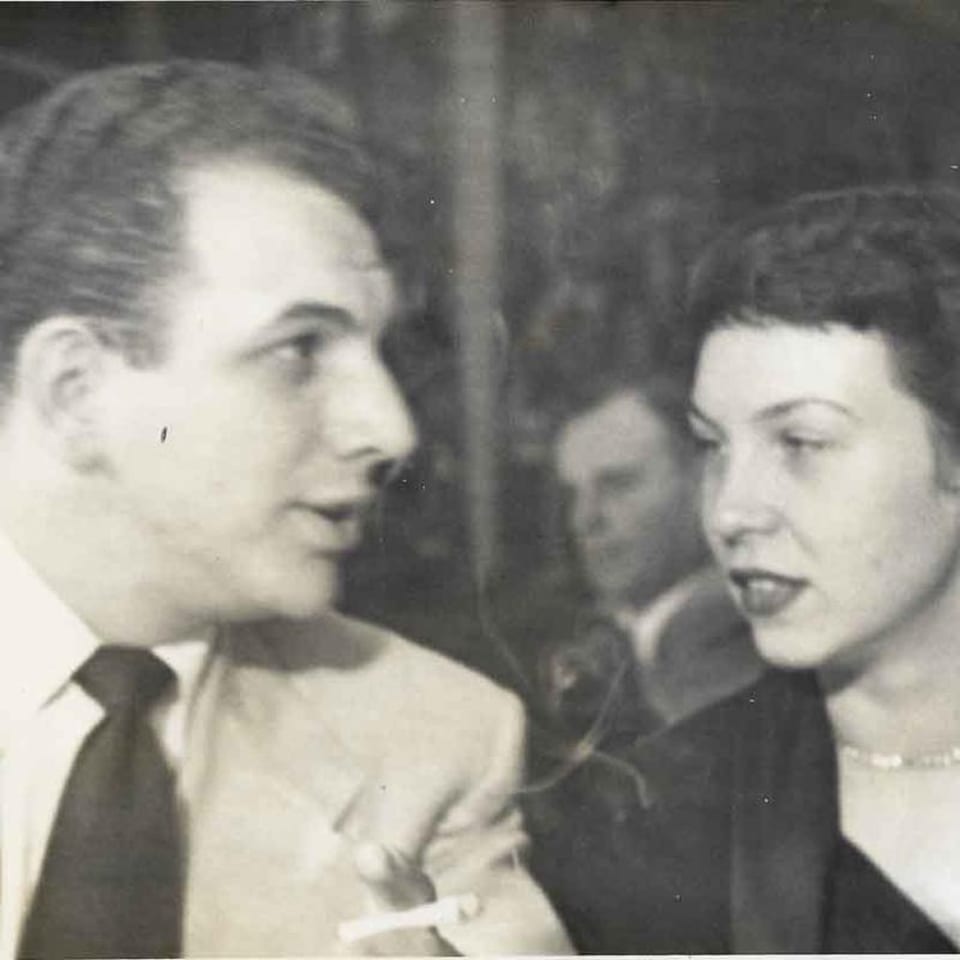 A vintage black and white photo of a man smoking and a woman talking, with another person in the background.