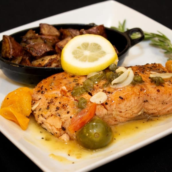 Salmon with lemon, capers, and garlic, served with roasted potatoes in a black dish.