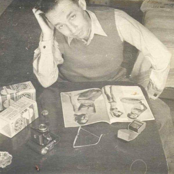A man in a vest and striped shirt sits at a table with a magazine, camera, and boxes.