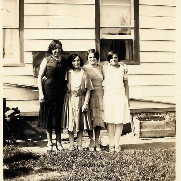 Four women in vintage dresses posing in front of a white clapboard house with windows.