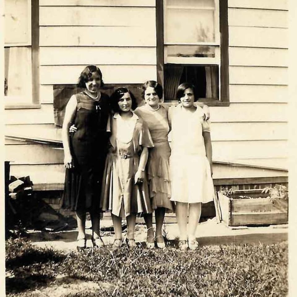 Four women in vintage dresses posing in front of a white clapboard house with windows.