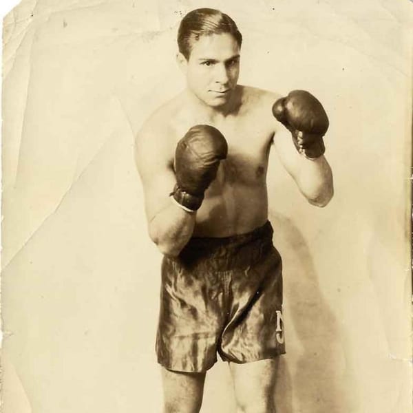 Vintage sepia photo of a shirtless male boxer in dark shorts and boxing gloves, ready to fight.