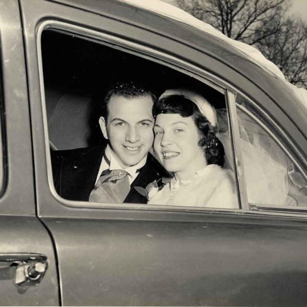 A smiling couple, possibly newlyweds, looking out from a car window in a vintage black and white photo.