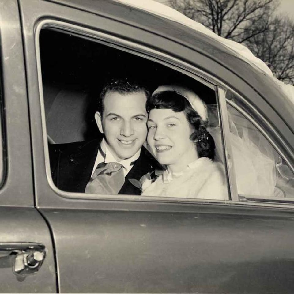 A smiling couple, possibly newlyweds, looking out from a car window in a vintage black and white photo.
