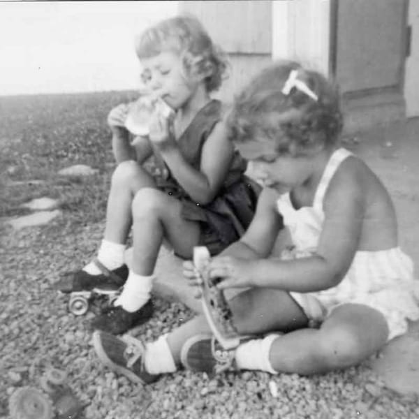 Two young girls, one on roller skates, sitting on a gravel path, playing outdoors.