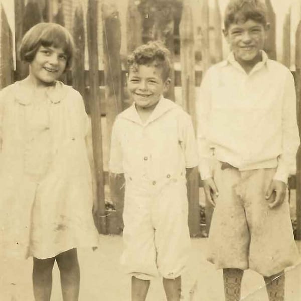 Three smiling children, two boys and a girl, stand together in an old sepia-toned photograph.