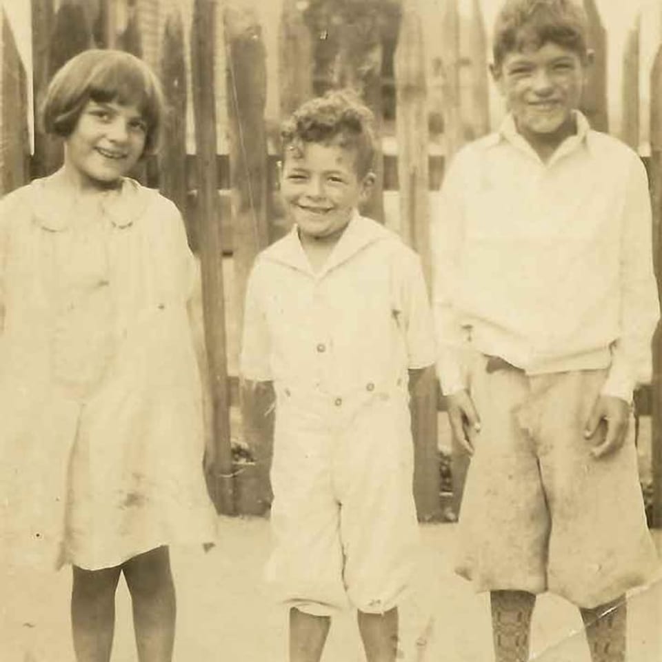 Three smiling children, two boys and a girl, stand together in an old sepia-toned photograph.